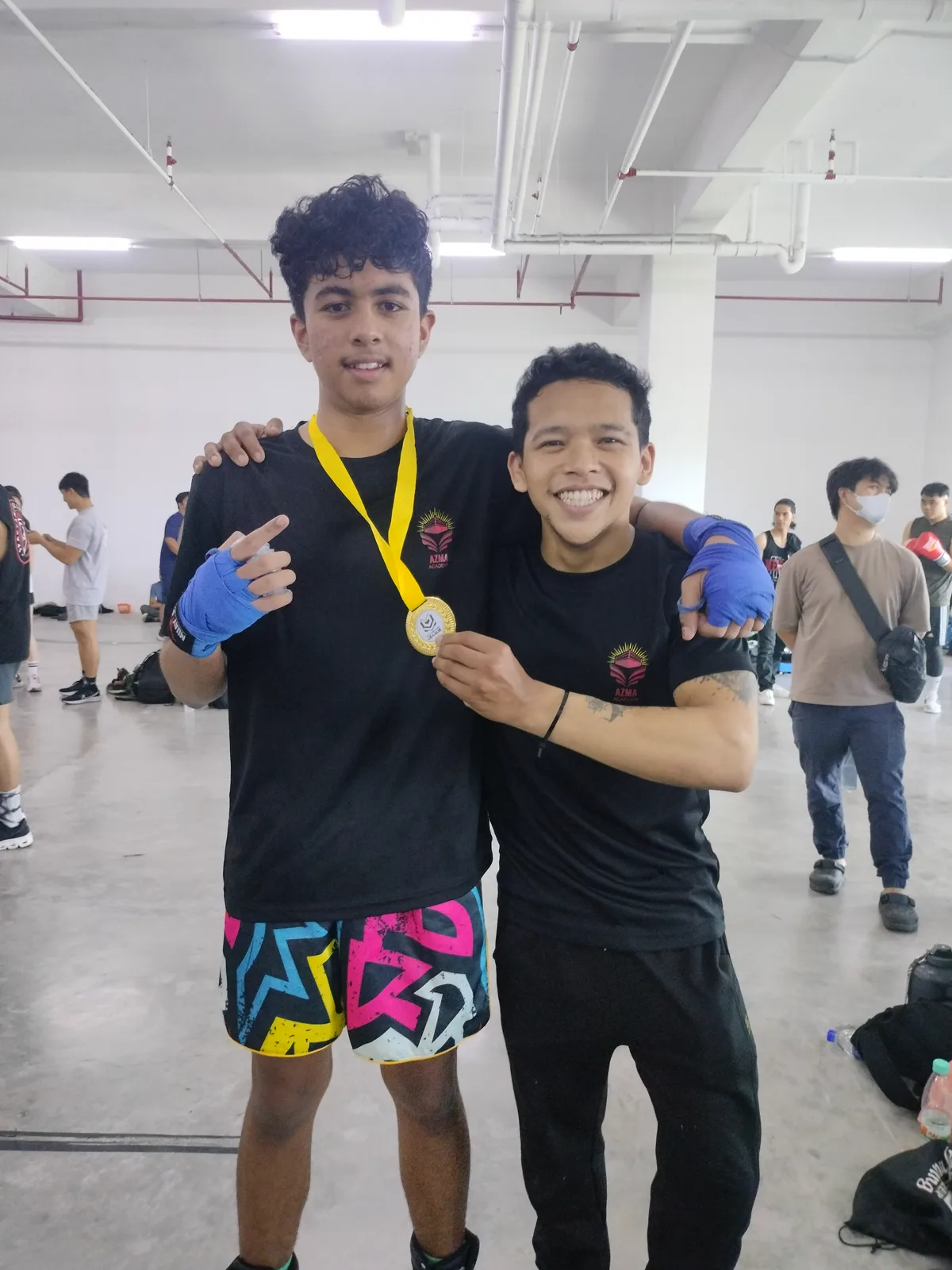 Coach Aiman and a smiling teen student in AZMA Academy shirts holding the student's gold medal